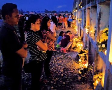 Filipinos stand in front of a wall of catacombs, lighting candles and offering food.